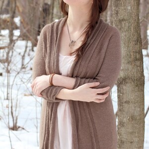 Puede incluir: Una mujer con un cárdigan marrón y una blusa rosa claro debajo. Está de pie en un bosque nevado con árboles en el fondo.