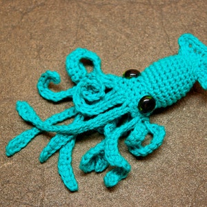 May include: A teal blue crocheted squid with green glass eyes. The squid has eight tentacles and a long, pointed body.