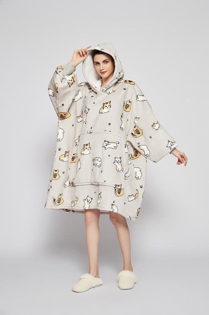 May include: A gray hooded blanket with a white fleece lining and a pattern of cartoon cats. The blanket has a large pocket in the front.