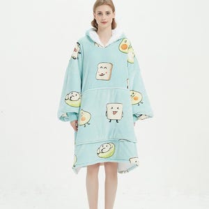 May include: A light blue hooded blanket with a pattern of smiling avocado and toast illustrations. The blanket has a large pocket in the front and is worn by a person.