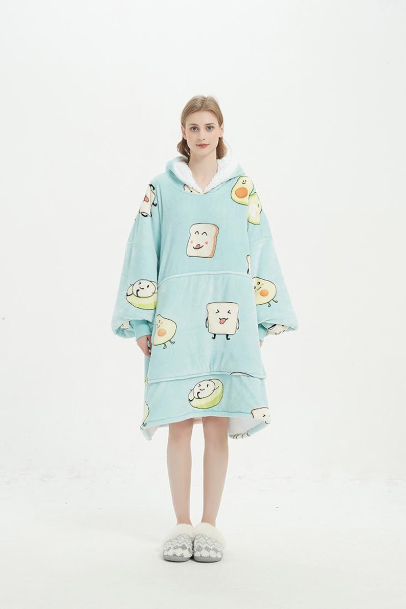 May include: A light blue hooded blanket with a pattern of smiling avocado and toast illustrations. The blanket has a large pocket in the front and is worn by a person.