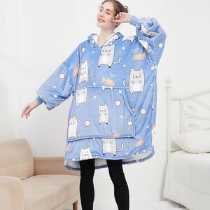 May include: A blue hooded blanket with a cat and fish pattern. The blanket has a large pocket on the front and is worn by a person.