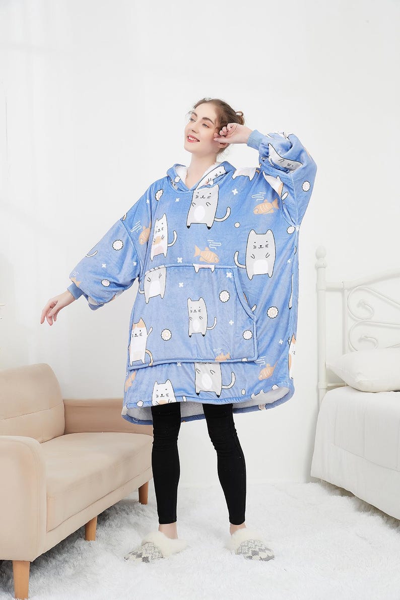 May include: A blue hooded blanket with a cat and fish pattern. The blanket has a large pocket on the front and is worn by a person.