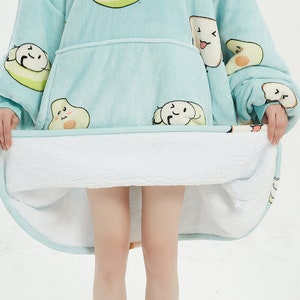 May include: A light blue hooded blanket with a white sherpa lining. The blanket features a cartoon avocado print with smiling faces.
