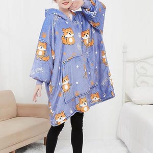 May include: A blue hooded blanket with a white and orange dog print. The blanket has a large pocket on the front and the words "Skipping Rope" and "Camping Trip" are printed on the sleeves.