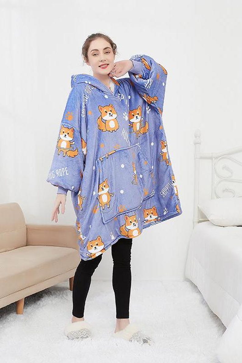 May include: A blue hooded blanket with a white and orange dog print. The blanket has a large pocket on the front and the words "Skipping Rope" and "Camping Trip" are printed on the sleeves.