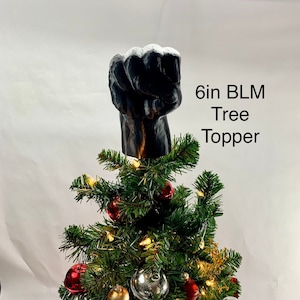 May include: A black 6-inch fist tree topper with white accents, placed atop a decorated Christmas tree. The tree features red, silver, and gold ornaments, and string lights. The text "6in BLM Tree Topper" is visible.