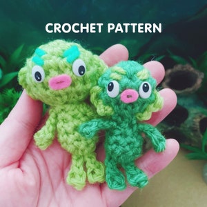 May include: Two green crocheted amigurumi figures with large eyes and pink mouths. The figures are in a hand, and the background is blurred.
