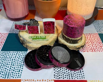 Puede incluir: Un conjunto de cuatro piezas de molinillo rosa y negro con un patrón de rosas, un encendedor blanco con un patrón de rosas y un molinillo rosa y negro con un patrón de rosas. Las piezas del molinillo están apiladas una encima de la otra. El encendedor está sentado sobre una base de madera. El molinillo está sentado sobre una base de madera. El texto "WISE FUME wisefume.com" está impreso en la base de madera.