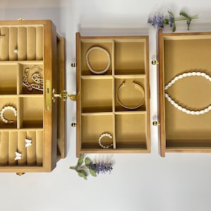 May include: A wooden jewelry box with multiple compartments, open to reveal various pieces of jewelry. The box is light brown with a gold clasp and hinges. Inside are rings, necklaces, bracelets, and earrings. A white pearl necklace is displayed in one compartment.