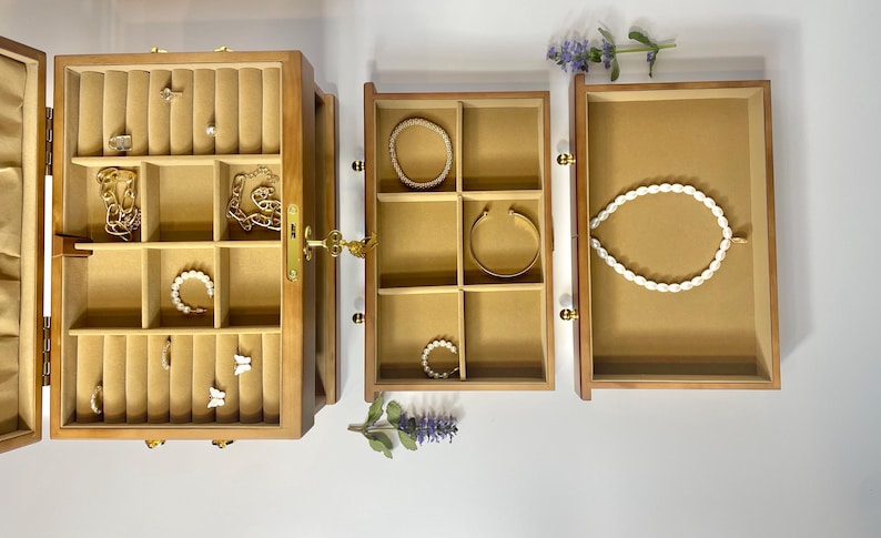 May include: A wooden jewelry box with multiple compartments, open to reveal various pieces of jewelry. The box is light brown with a gold clasp and hinges. Inside are rings, necklaces, bracelets, and earrings. A white pearl necklace is displayed in one compartment.
