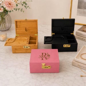 Puede incluir: Tres cajas decorativas: una caja de bambú marrón claro, una caja negra y una caja rosa. Las cajas tienen compartimentos, una cerradura de combinación y una tapa con bisagras. La caja rosa tiene el nombre "Rachel" y un diseño floral.