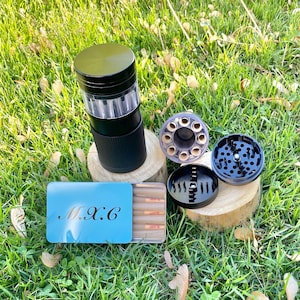 May include: A black and silver herb grinder with a clear chamber, disassembled next to a blue metal box with the letters "M.X.C" and pre-rolled cones. The items are on a bed of green grass.