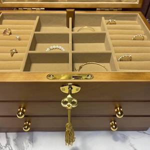 May include: A wooden jewelry box with multiple compartments lined with beige fabric. The box is open, revealing various rings, bracelets, and necklaces. The box has a gold key and tassel, and gold knobs on the drawers.