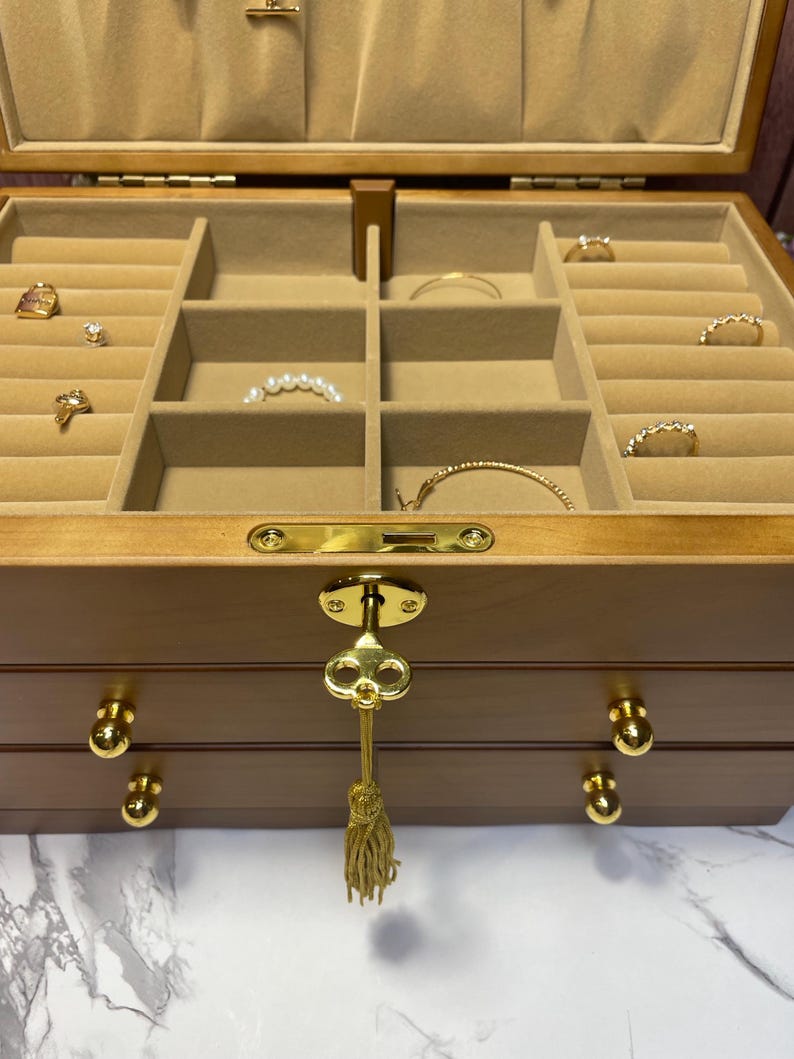 May include: A wooden jewelry box with multiple compartments lined with beige fabric. The box is open, revealing various rings, bracelets, and necklaces. The box has a gold key and tassel, and gold knobs on the drawers.