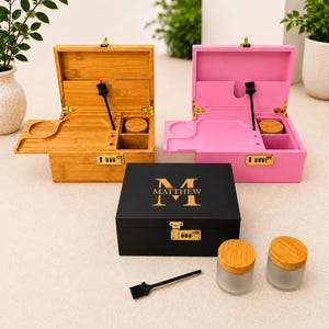 May include: Three wooden boxes, one bamboo-colored, one pink, and one black, are displayed open and closed. The black box has "M MATTHEW" in gold lettering. Each box has a combination lock and a small tool. Two glass jars with wooden lids are also present.