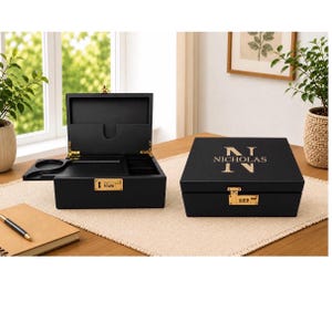May include: Two black wooden boxes with gold accents. One box is open, revealing compartments and a tray. The other box is closed, with the name "NICHOLAS" and the initial "N" printed in gold. Both boxes have combination locks.