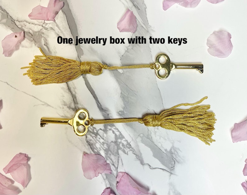 May include: Two gold-colored keys with decorative tassels, lying on a marble surface. The keys are connected to gold tassels. The text "One jewelry box with two keys" is visible.