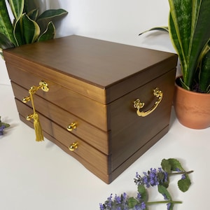 May include: A wooden jewelry box with a warm brown finish. It features three drawers with gold-colored knobs and a gold-colored handle. A gold-colored tassel hangs from the keyhole. The box is rectangular and sits on a white surface.
