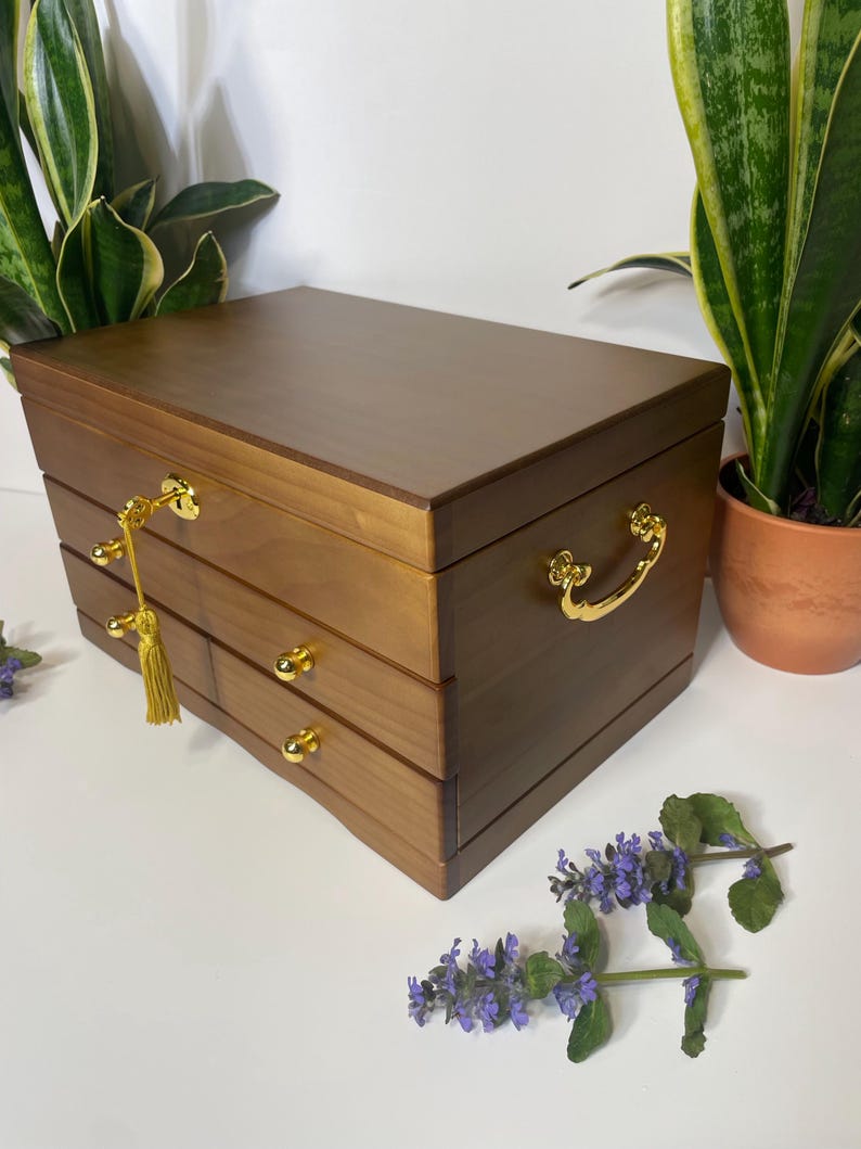 May include: A wooden jewelry box with a warm brown finish. It features three drawers with gold-colored knobs and a gold-colored handle. A gold-colored tassel hangs from the keyhole. The box is rectangular and sits on a white surface.