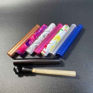 Puede incluir: Un conjunto de siete tubos metálicos coloridos y con patrones, con un tubo negro y un papel de liar blanco. Los tubos tienen diferentes diseños, incluyendo dinero, flores y patrones abstractos.