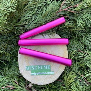 May include: Three bright pink cylindrical objects rest on a wooden slice, surrounded by green foliage. The objects are evenly spaced and appear to be made of metal. A green sticker with the text "WISE FUME" and the website "wisefume.com" is visible.