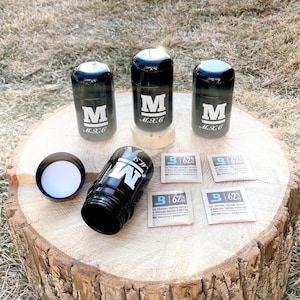 Puede incluir: Cuatro tarros de vidrio negro con una letra blanca "M" y el texto "M.X.C" en la parte delantera. Un tarro está abierto y la tapa está al lado. Hay cuatro pequeños paquetes de papel marrón con el texto "B 62%" en ellos.