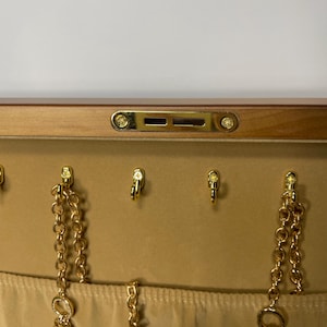 May include: A jewelry box with a light brown wooden exterior and a beige interior. The interior features gold-colored hooks and chains with charms, including a key, a lock, and a cross. The box has a gold-colored clasp.