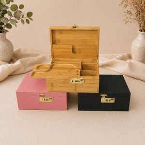 Könnte beinhalten: Drei Holzkisten: eine offene, eine rosa und eine schwarze. Die offene Kiste ist aus Bambus mit Fächern und einem eingebauten Tablett. Die rosa und schwarzen Kisten sind mit Zahlenschlössern verschlossen. Die Kisten stehen auf einer neutralen Oberfläche.