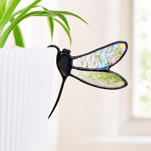 May include: A dragonfly-shaped plant stake featuring a black metal frame and iridescent stained glass wings. The wings display a textured, shimmering surface with hints of green, blue, and white tones, attached to a thin black metal stem.
