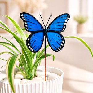May include: A blue butterfly garden stake with black outlines and white accents, placed in a white pot with a green plant. The butterfly has detailed wing patterns and is made of metal.