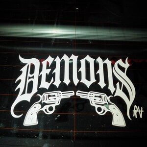 Könnte beinhalten: Weißer Aufkleber mit dem Wort "DEMONS" in gotischer Schrift, flankiert von zwei Revolvern. Der Aufkleber befindet sich auf einer schwarzen Oberfläche, möglicherweise einer Autoscheibe. Enthält die Initialen "AV".