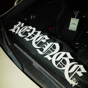 May include: White vinyl decal with the word "REBEL" in a gothic font, applied to a car windshield. The decal is centered and spans the width of the glass. An air freshener hangs from the rearview mirror.
