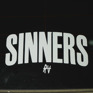 Puede incluir: Texto blanco "SINNERS" con un logotipo "AV" estilizado debajo, sobre un fondo negro. El texto es en negrita, sin serif, creando un diseño gráfico y llamativo.