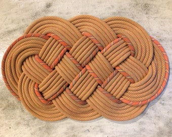 May include: A large, oval-shaped rug made from woven rope. The rope is a light brown color with orange and blue accents. The rug is knotted in a complex pattern.