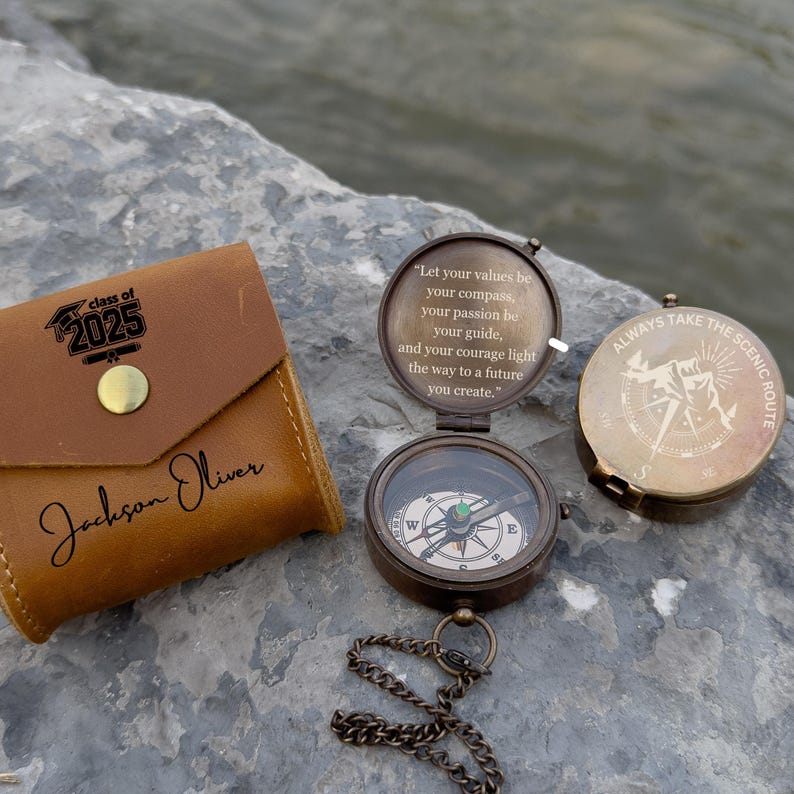 May include: A collection of compasses and a leather wallet. The open compass displays a green and white directional dial. The wallet is brown with the text "Class of 2025" and a name. One compass reads "Always take the scenic route."