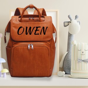 K&ouml;nnte beinhalten: Ein brauner Leder-Wickeltasche-Rucksack mit dem Namen "OWEN" in Schwarz auf der Vorderseite.