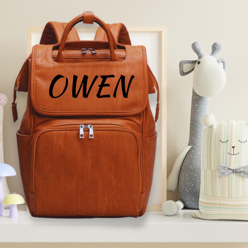 K&ouml;nnte beinhalten: Ein brauner Leder-Wickeltasche-Rucksack mit dem Namen "OWEN" in Schwarz auf der Vorderseite.