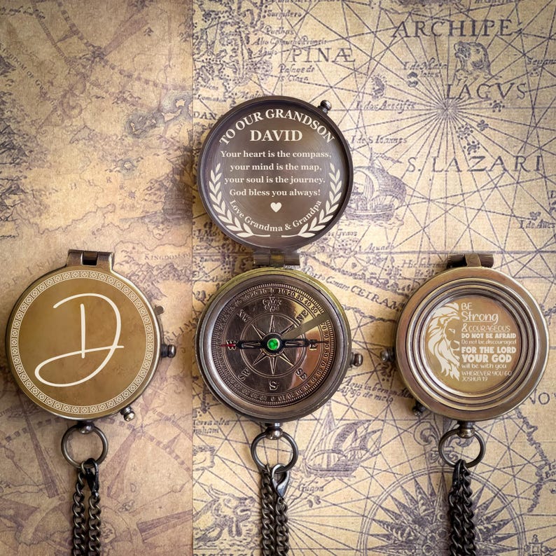 May include: Three antique-style compasses with detailed designs and chains. One compass is open, displaying text that reads "TO OUR GRANDSON DAVID." The other two compasses have decorative initials and inspirational quotes. The background is a vintage map.