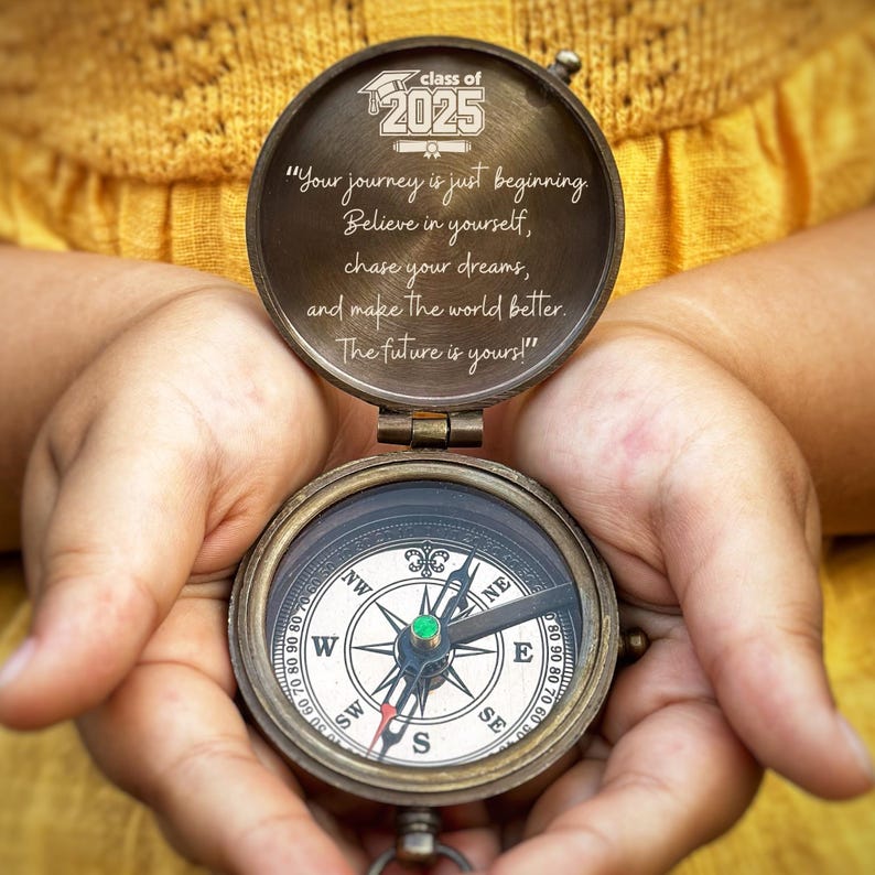 May include: A vintage brass compass with the lid open, held in hands. The lid reads "Class of 2025" and an inspirational quote. The compass face shows directional markings. The background is a blurred yellow.