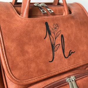 K&ouml;nnte beinhalten: Ein brauner Lederrucksack mit Rei&szlig;verschluss und einem floralen Monogramm, das "Mia" sagt.