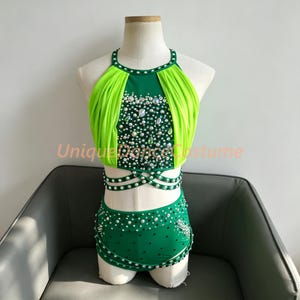 May include: A two-piece dance costume featuring a bright green halter top with pleated lime green side panels and a matching high-waisted bottom. Both pieces are heavily embellished with clear crystals and beads. The text UniqueDanceCostume is visible.