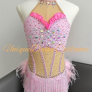 May include: A light pink dance costume featuring a high neck, sheer mesh panels, and a bodice adorned with numerous crystals and rhinestones. The garment includes a ruffled neckline, fringe detailing at the waist, and light pink feather accents.