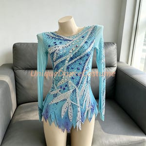 Può includere: Un body a maniche lunghe color azzurro con un complesso motivo di cristalli argentati e blu scuro disposti in linee vorticose. Il capo presenta uno scollo rotondo, maniche in rete trasparente e un orlo frastagliato e appuntito.