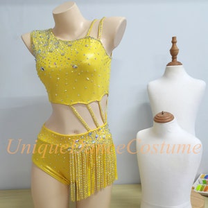 以下が含まれることがあります： ラインストーンの装飾が施された黄色のダンスコスチューム。片方の肩にストラップが付いたクロップトップとカットアウトデザイン、ショートパンツとフリンジスカートが特徴です。マネキンに着せられています。