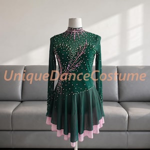 May include: A dark green long-sleeve dance costume featuring a high neckline, sheer mesh fabric, and a layered skirt with a light pink underskirt. The bodice is embellished with scattered white crystals and pink floral-like patterns. UniqueDanceCostume.