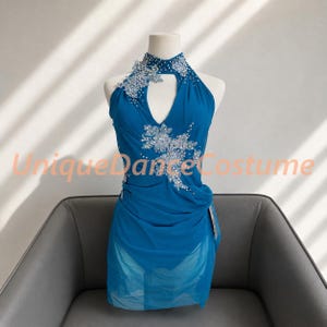 May include: A bright blue sleeveless dance costume featuring a high neckline with a teardrop cutout. The bodice is adorned with silver floral-shaped crystal embellishments. The skirt is made of layered sheer fabric with a gathered, draped design.