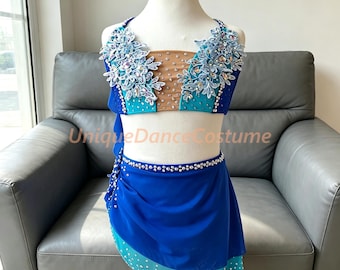 May include: A two-piece dance costume featuring a royal blue and turquoise top with nude mesh paneling, adorned with white floral lace appliques and clear crystals. The matching royal blue skirt has a layered hem and crystal embellishments.