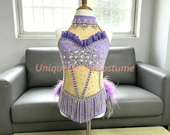 Puede incluir: Un traje de baile color lavanda con cuello alto, corpiño con volantes y paneles de malla transparente en tono nude. La prenda está decorada con cristales transparentes y lentejuelas plateadas, con plumas moradas en las caderas y flecos.