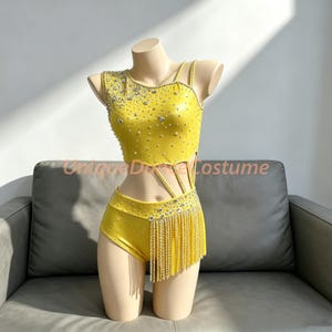May include: A bright yellow two-piece dance costume featuring a sleeveless crop top and high-waisted shorts. The set is adorned with scattered clear rhinestones and features a side panel of long, shimmering yellow fringe on the shorts.